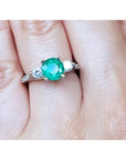 Round cut emerald and two side diamonds in a 14K white gold three-stone ring on a finger.