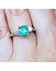 Round cut emerald and two side diamonds in a 14K white gold three-stone ring on a finger.