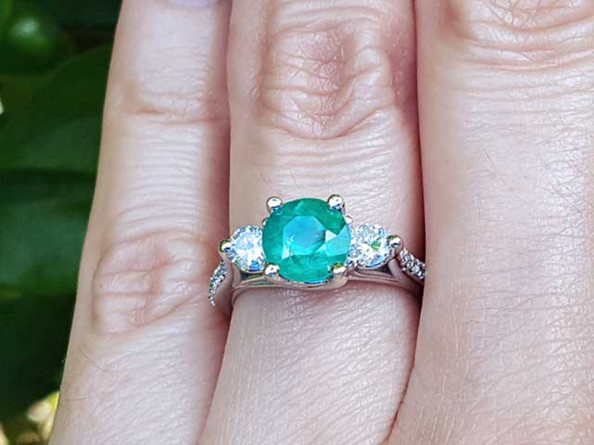 Emerald ring with side natural diamonds on a finger with a blurred green background.