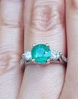 Emerald ring with side natural diamonds on a finger with a blurred green background.