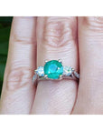 Emerald ring with side natural diamonds on a finger with a blurred green background.