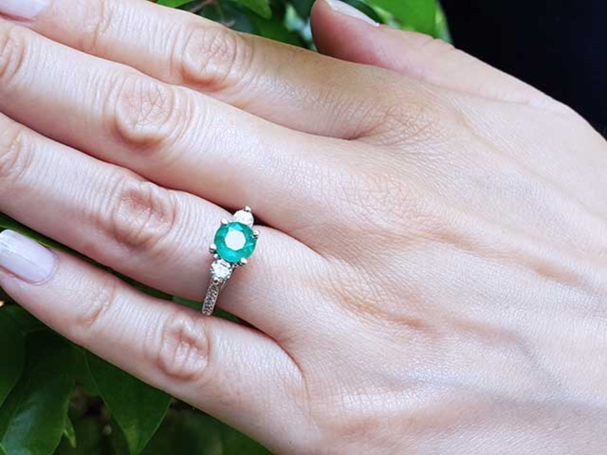 Hand wearing a ring with an emerald stone against a green leafy background.