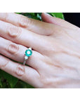 Hand wearing a ring with an emerald stone against a green leafy background.