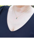 Petite round cut deep green Colombian emerald necklace with halo diamonds in 14k yellow gold on a woman's neck.