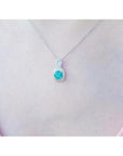 Round Colombian emerald and diamond halo pendant in 18K white gold, worn on a woman's neck.