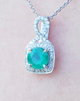 A 18k white gold pendant with a natural colombian emerald on a light pink background.