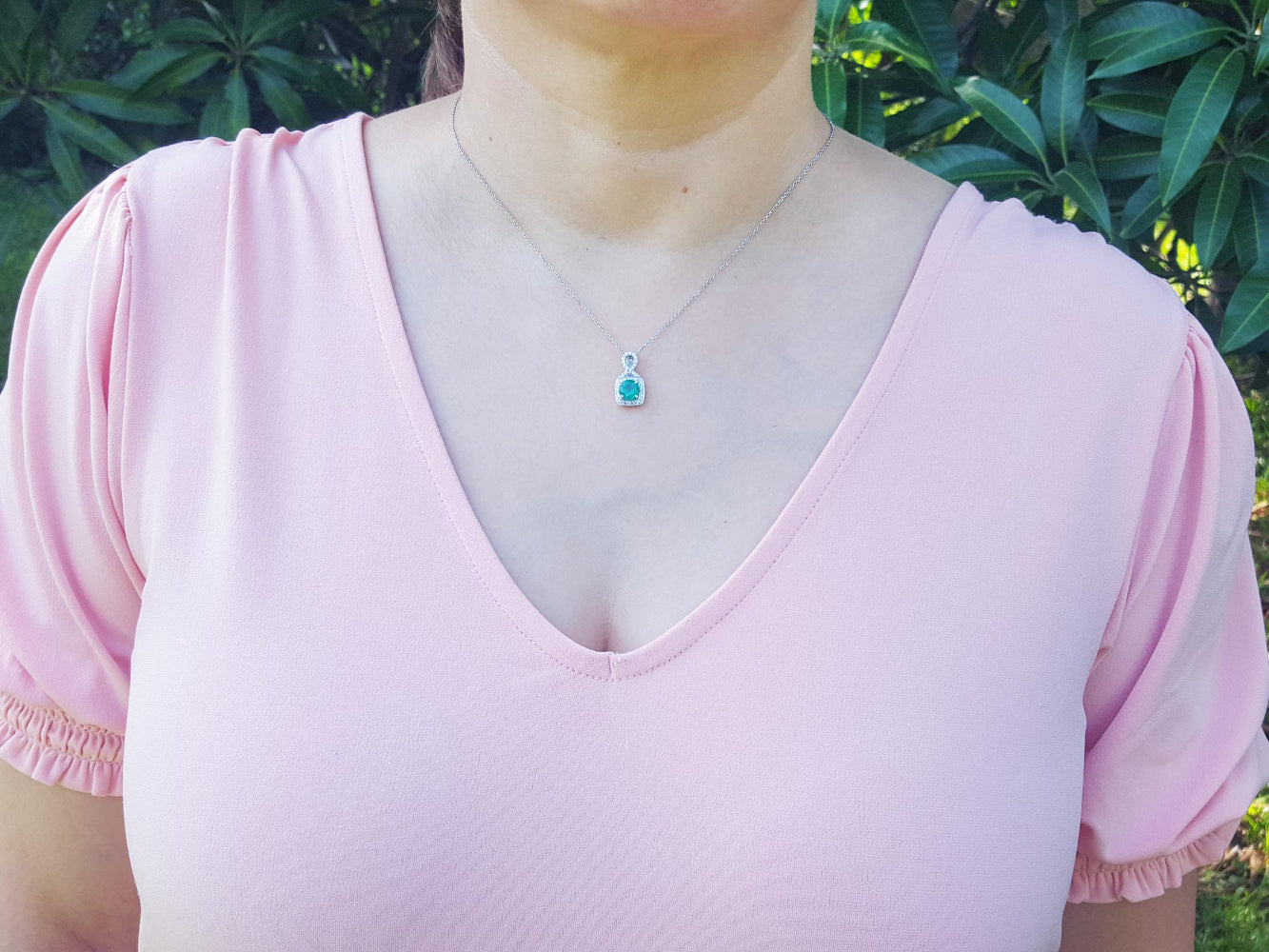A woman wearing a pink top with a natural emerald pendant necklace against a green leafy background