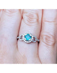 A round cut natural emerald surrounded by a halo diamond in 14k white gold on a finger.