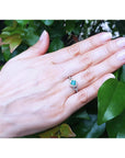 Hand wearing a ring with a round cut emerald against a leafy background.