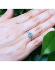 Hand wearing a white gold ring with a round cut emerald against a green leafy background.