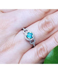 14K white gold ring with a round bluish-green emerald haloed by diamonds on a finger.