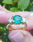 A 18k yellow gold ring with a round cut emerald held between fingers against a blurred green leafy background.