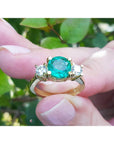 A 18k yellow gold ring with a round cut emerald held between fingers against a blurred green leafy background.