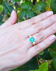 Hand wearing a round cut emerald and side diamonds ring against a green leafy background.