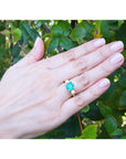 Hand wearing a round cut emerald and side diamonds ring against a green leafy background.
