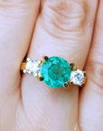 18k yellow gold round cut emerald ring with two side diamonds on a finger.