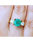 18k yellow gold round cut emerald ring with two side diamonds on a finger.