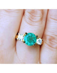 18k yellow gold GIA certified round cut colombian emerald ring with two side diamonds on a finger.