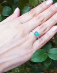 Hand wearing a ring with an emerald stone against a green leafy background.