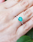 Hand wearing a white gold ring with a round cut emerald against a natural background.