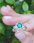 Hand holding a brides' emerald engagement rings against a green leafy background.