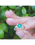 Hand holding a brides' emerald engagement rings against a green leafy background.
