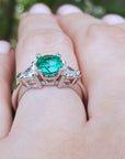 Emerald ring with diamond accents on a hand against a blurred green background