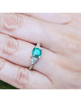 1.00 ct. round emerald ring with trillion side diamonds on a 14K white gold band, worn on a finger.