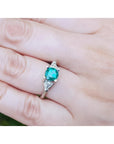 1.00 ct. round emerald ring with trillion side diamonds on a 14K white gold band, worn on a finger.