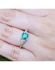 1.00 ct. round emerald ring with trillion side diamonds on a 14K white gold band, worn on a finger.