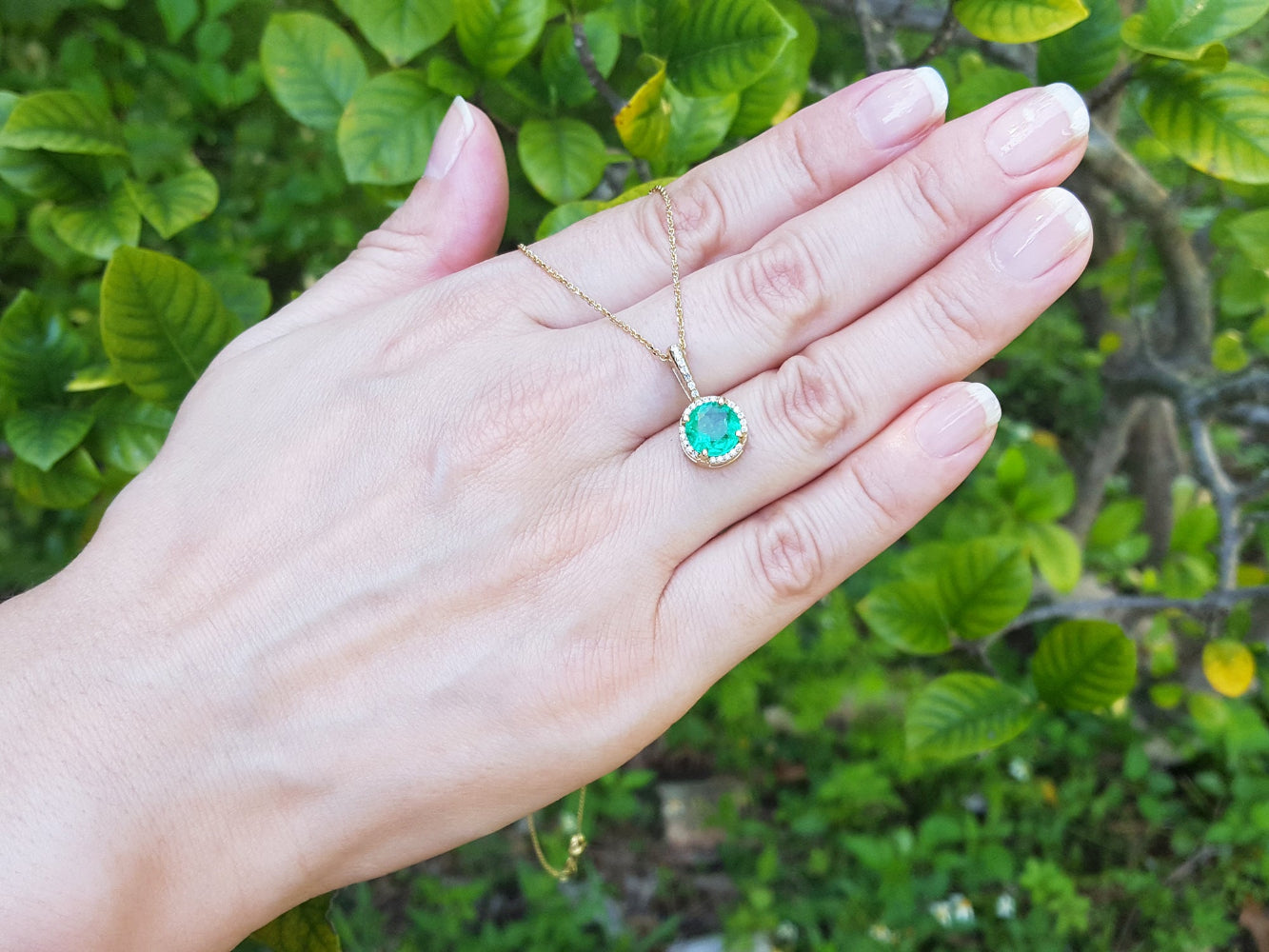 Hand displaying a gold pendant with a natural emerald against a green leafy background.