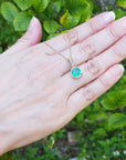 Hand displaying a gold pendant with a natural emerald against a green leafy background.