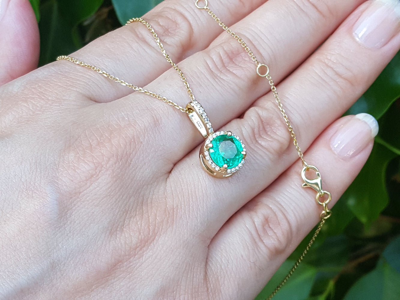 Gold necklace with a colombian emerald pendant on a hand against a green leafy background.