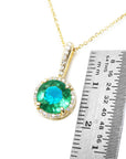 Gold necklace with a round cut emerald pendant next to a ruler for scale on a white background.