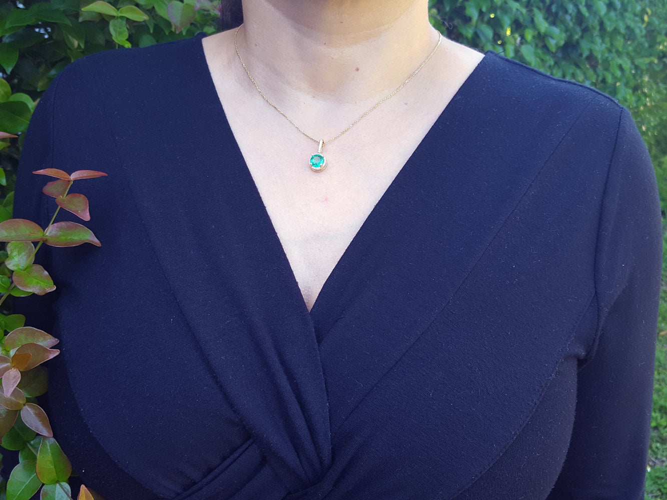 A woman wearing a navy-blue dress with an emerald pendant necklace against a green leafy background.