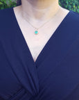 A woman wearing a navy-blue dress with an emerald pendant necklace against a green leafy background.