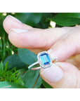 14k white gold ring with a blue square natural sapphire held between fingers against a green leafy background