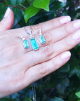 A hand displaying a trio set of emerald earrings and pendant against a green leafy background