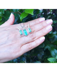 A hand displaying a trio set of emerald earrings and pendant against a green leafy background