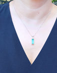 A woman wearing a blue top with a necklace featuring a rectangular emerald pendant.