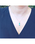 A woman wearing a blue top with a necklace featuring a rectangular emerald pendant.