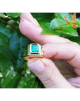 Gold ring with a real emerald held between fingers against a leafy background