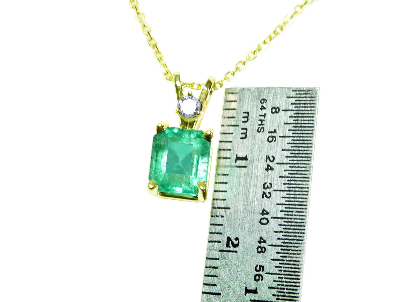Gold pendant with a natural emerald-cut emerald next to a ruler for size reference on a white background.