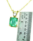 Gold pendant with a natural emerald-cut emerald next to a ruler for size reference on a white background.