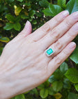 Hand wearing an emerald ring with a green gemstone against a green leafy background.