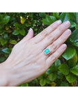 Hand wearing an emerald ring with a green gemstone against a green leafy background.