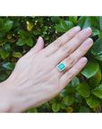 Emerald-cut bluish-green emerald ring with halo diamonds in 14K white gold on a hand.