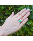 Emerald-cut bluish-green emerald ring with halo diamonds in 14K white gold on a hand.