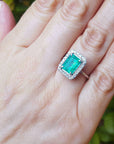 Hand wearing a square emerald ring with a halo diamond against a green leafy background.