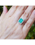 Emerald-cut bluish-green emerald ring with halo diamonds in 14K white gold on a woman's hand.
